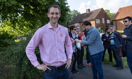 Andrew Lewin canvassing: he wears a pink shirt and stands smiling in front of a hedge on a housing estate while fellow party workers gather behind him.